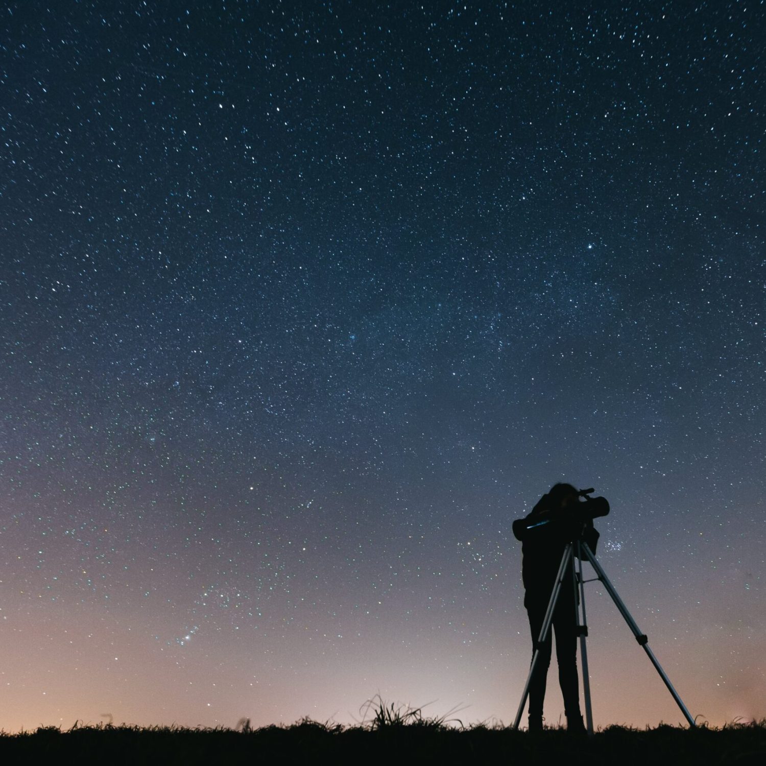 A silhouetted person using a telescope to observe the night sky filled with stars.